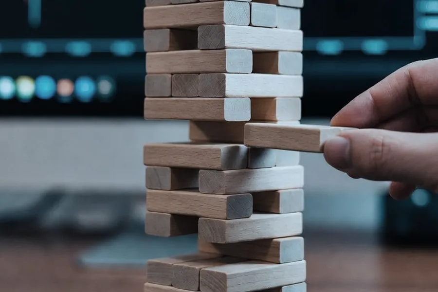 A hand carefully pulls one wooden block from the middle of a tall block tower, with a blurred trading chart on a monitor behind it